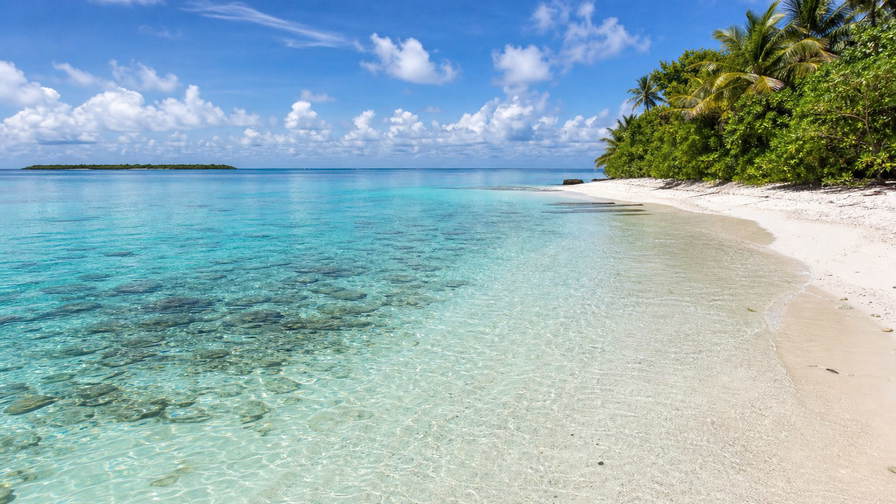 Brits Indische Oceaanterritorium: zonovergoten atolrand met glashelder turquoise lagunewater, krijtwit koraalzand in de ondiepten dat rifschaduw vlecht onder passaat-cumulus en kokospalmen langs de zoom in de hero-foto - visuele setting voor onafhankelijke vergelijkingen van kosten van leven, supermarkt- en horecaprijzen rond afgelegen Britse overzeese gebieden.