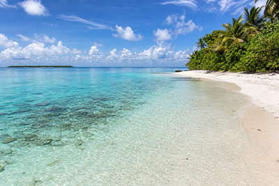 Brits Indische Oceaanterritorium: zonovergoten atolrand met glashelder turquoise lagunewater, krijtwit koraalzand in de ondiepten dat rifschaduw vlecht onder passaat-cumulus en kokospalmen langs de zoom in de hero-foto - visuele setting voor onafhankelijke vergelijkingen van kosten van leven, supermarkt- en horecaprijzen rond afgelegen Britse overzeese gebieden.