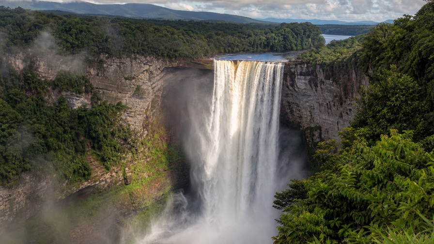 Guyana: waterval Kaieteur met zandstenen klif en jadekleurige kloof die gouden nevel onder handels-cumulus vlecht, nevel boven het regenwoudcanopy van Iwokrama door koperkleurige glans op de Essequibo naar pastelkleurige arcade-stapels van Stabroek Market in Georgetown en stofbanden boven termietenheuvels in de savanne van Rupununi in de hero - achtergrond voor marktbriefjes in Guyaanse dollar (GYD), gevouwen USD-rondvlucht-charter-stempels bij ijskades op vliegveld Ogle en weegbriefjes voor cashewnoten in Lethem.