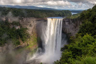 Guyana: waterval Kaieteur met zandstenen klif en jadekleurige kloof die gouden nevel onder handels-cumulus vlecht, nevel boven het regenwoudcanopy van Iwokrama door koperkleurige glans op de Essequibo naar pastelkleurige arcade-stapels van Stabroek Market in Georgetown en stofbanden boven termietenheuvels in de savanne van Rupununi in de hero - achtergrond voor marktbriefjes in Guyaanse dollar (GYD), gevouwen USD-rondvlucht-charter-stempels bij ijskades op vliegveld Ogle en weegbriefjes voor cashewnoten in Lethem.