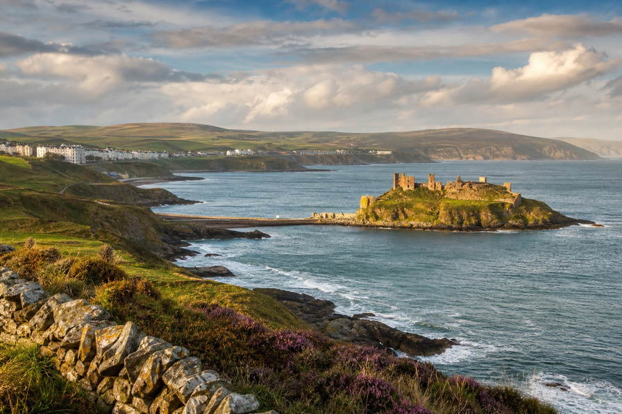 Isle of Man: graniet-vlecht van de promenade van Douglas over haven-chopbanden van de Ierse Zee onder handels-cumulus, kopernevel van het Snaefell-moor naar perkamentstapels bij Peel Castle en lint van Laxey-glen bij het waterrad Lady Isabella plus weidegruis van Manx Loaghtan-schapen in de hero - achtergrond voor GBP markt-streepjes met Manx-pond-sterling-pariteit-stroken, EUR Ierse-Zee-veerboot-charter-stempelvouwen bij ponton-stapels van Douglas Sea Terminal en rook-haring-weegbriefjesbanden in Ramsey-baai.