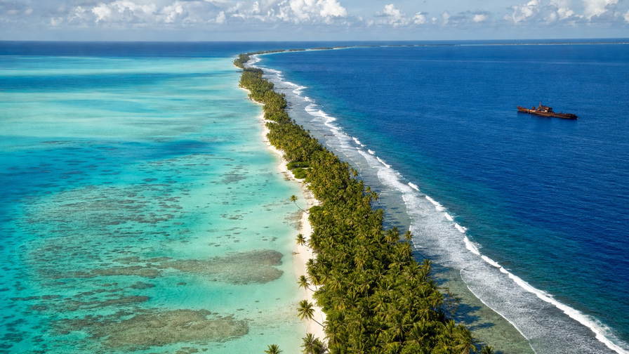 Kiribati: turquoise atolband rond Tarawa die dubbele spiegel van oceaan en lagune vlecht onder passaat-stratus, roestkleurige wraksilhouetten bij Betio naar geweven mattenstallen in Bairiki kopra-zakpiramides en zilveren strepen van noddy-vlucht boven het rif in de hero - achtergrond voor marktzettels in Australische dollars (AUD), diesel-buitenboordliterbonnen en weegbriefjes voor kopra naar de buiten-eilanden.