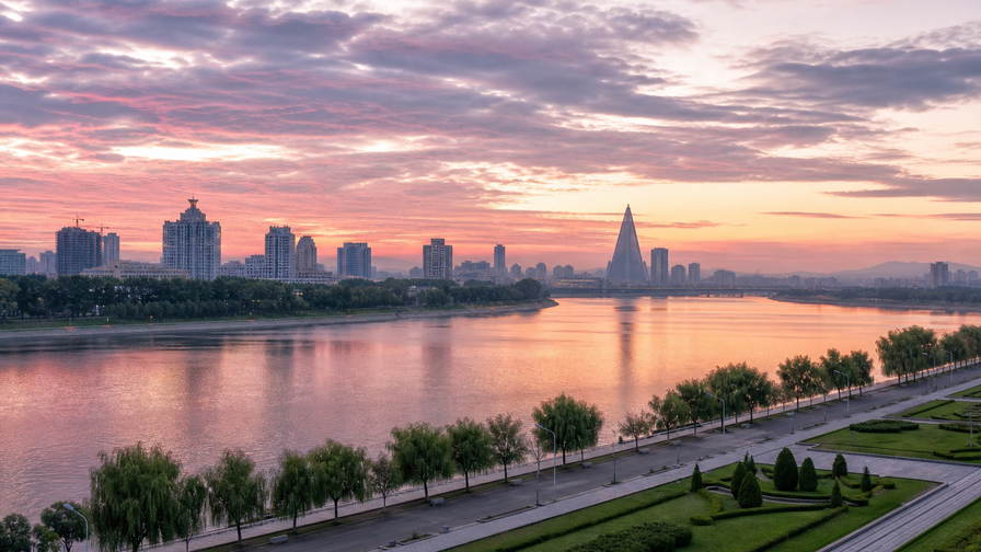 Noord-Korea: Taedong-oever-esplanade-vlecht over dageraad-chopbanden onder stratocumulus, kopernevel van skyline-silhouet naar perkamentstapels bij Mansu-heuvel en bassinlint van fontein bij haven-ponton-groeven plus dennenpollen op helling in de hero - achtergrond voor EUR-geïndexeerde markt-streepjes met KPW-contant-echo, USD logistiek-charter-stempelvouwen bij glijkais van Nampo en Taedong-vis-weegbriefjesbanden.