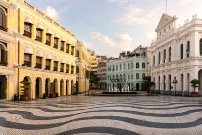 Macau: zwart-wit golfpatroon van het plaveisel op het Senado-plein dat pasteltinten van neoklassieke arcadegevels vlecht onder moessoncumulus, stenen façade van de ruïne van St. Paul naar LED-glans langs de Cotai-kust en rode luiken in Rua da Felicidade met witte schijf van vuurtoren Guia in de hero - achtergrond voor briefjes van pastéis de nata-kraampjes in patacas (MOP), gevouwen stempels van de veerboot in de Buitenhaven en bonnetjes voor stomende broodjes met varkenskotelet.