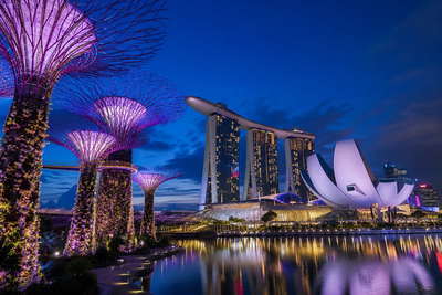 Singapore: verticale supertree-tuinen in Marina Bay die paarse lasernevel van lotusblaadjes van ArtScience Museum vlechten, neonrimpelingen bij Clarke Quay richting satérook van hawkercentra onder wasgoed-geometrie aan HDB-flats en robijnrode avondstrepen van de kabelbaan naar Sentosa in de hero - achtergrond voor jetons bij hawkerkramen in Singaporese dollar (SGD), gevouwen bonnen voor kopi-o met sokfilter en stapels EZ-link-opwaardeerstrookjes.