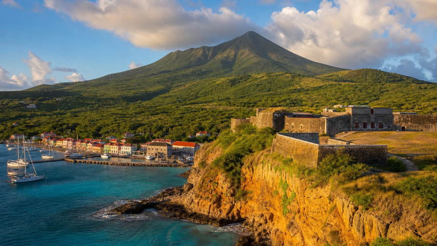 Sint Eustatius: kamienny splot dolnego Oranjestad sznurem karaibskiej fali pod kumulusami, miedziana mgielka Fort Oranje ku pergaminowym stosom kaldery The Quill i wstega pasa lotniska Golden Rock przy pontonach lodzi nurkowych oraz pyl hibiskusa na zboczu w obrazie - tlo dla zapisow XCD na targu, USD stempli czarteru jachtowego przy slipach Gallows Bay i tasm wazenia ryb na rynku przy dawnych solankach.