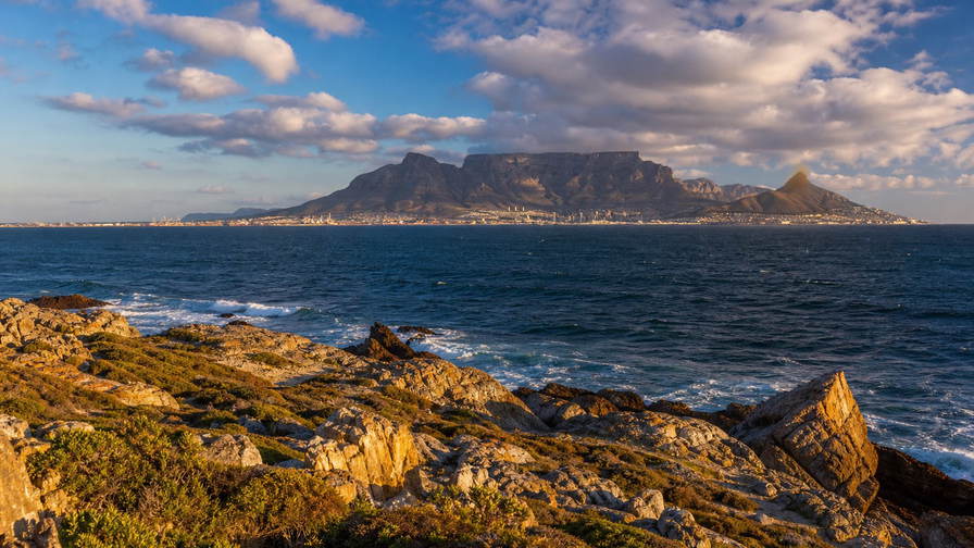 Zuid-Afrika: tafelberg-plateau-skyline boven de Atlantische Oceaan bij warm laag licht, fynbos en rotsachtige Kaapse voorgrond die overloopt in baai-deinigingsbanden onder mooiweer-stapelwolken in de hero-foto - visueel anker voor onafhankelijke supermarkt-, wijnland-horeca- en stadhuurprijsvergelijkingen.