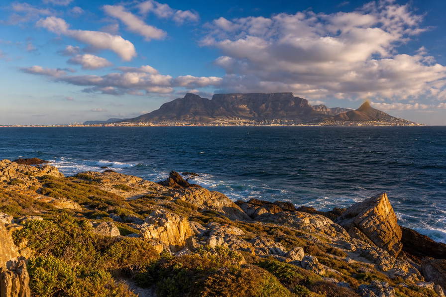 Zuid-Afrika: tafelberg-plateau-skyline boven de Atlantische Oceaan bij warm laag licht, fynbos en rotsachtige Kaapse voorgrond die overloopt in baai-deinigingsbanden onder mooiweer-stapelwolken in de hero-foto - visueel anker voor onafhankelijke supermarkt-, wijnland-horeca- en stadhuurprijsvergelijkingen.