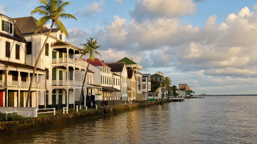 Suriname: pastelkleurige houten arcade aan de Waterkant die jadekleurige branding van de Surinamerivier onder handels-cumulus vlecht, nevel aan de kam van Brownsberg door regenwoud naar kalkstenen wallen van fort Nieuw Amsterdam en koperkleurige nevelbanden boven het Brokopondo-reservoir met perkselstof van sloepen op de Commewijne in de hero - achtergrond voor kadebriefjes in Surinaamse dollar (SRD), gevouwen USD-charter-stempels bij ijskades van jachthaven Marina Suriname en weegbriefjes voor vis op de grensmarkt in Albina.