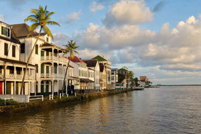 Suriname: pastelkleurige houten arcade aan de Waterkant die jadekleurige branding van de Surinamerivier onder handels-cumulus vlecht, nevel aan de kam van Brownsberg door regenwoud naar kalkstenen wallen van fort Nieuw Amsterdam en koperkleurige nevelbanden boven het Brokopondo-reservoir met perkselstof van sloepen op de Commewijne in de hero - achtergrond voor kadebriefjes in Surinaamse dollar (SRD), gevouwen USD-charter-stempels bij ijskades van jachthaven Marina Suriname en weegbriefjes voor vis op de grensmarkt in Albina.