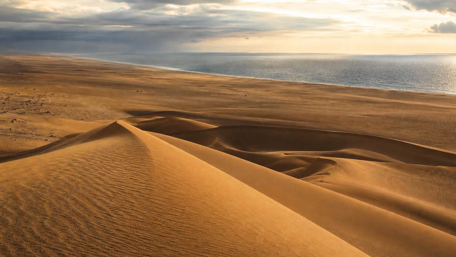 Westelijke Sahara: Atlantische duinenkam vlecht Laayoune golfbanden onder handelsstapelwolken, koper-jade nevel van Dakhla-lagune naar Smara-plateau en perkamentstapels aan de kade, erglint in kustmist-haar plus stof van de weg naar Aousserd in de hero - achtergrond voor MAD souk-streepjes, EUR veerboot-charter-stempelvouwen bij havenglijkais in Dakhla en USD sardien-weegbriefjesbanden.