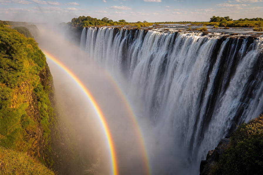 Zimbabwe: regenboog in de spray van de Victoriawatervallen boven de basalt-rand van Batoka-kloof aan de Zambezi, droge stenen kegelmuur van Great Zimbabwe in avondgoud en lantaarns op de Avondale-weekendmarkt in Harare in de hero - achtergrond voor sadza met relish, zeepsteen-kunst in Victoria Falls en cafébonnen in Amerikaanse dollars met mobiele-wallet-opwaarderingen.